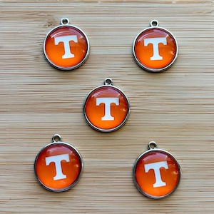 May include: Five round charms with a silver-colored border. Each charm features a white letter "T" on an orange background. The charms are arranged on a light brown wooden surface.