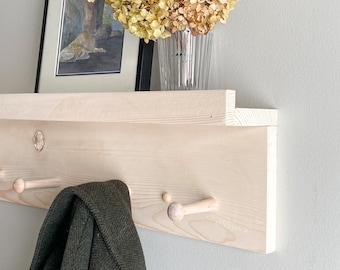 May include: A light-colored wooden wall-mounted shelf featuring a horizontal ledge and multiple protruding pegs for hanging items. A dark green coat hangs from one peg, while a framed picture and a glass vase with dried flowers sit on top.