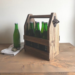 Peut inclure: Porte-bière en bois pour six bouteilles avec un ouvre-bouteille. Le porte-bière est de couleur brune et a un aspect rustique. Il contient cinq bouteilles de bière en verre vert.