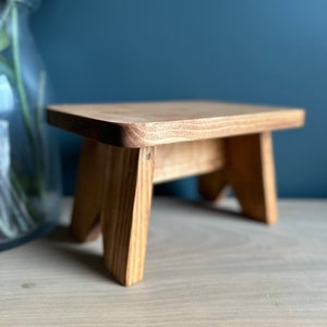 May include: A small wooden stool with a natural wood finish. The stool has a rectangular top and four legs.