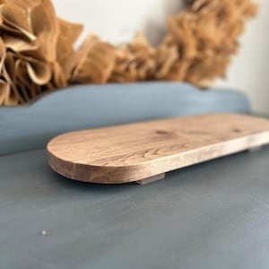 May include: A light brown wooden riser with a smooth, oval-shaped top surface and rounded edges. The piece features a natural wood grain pattern and is supported by small, low-profile feet that elevate the platform slightly above the surface.