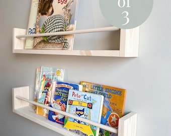 May include: A set of three light-colored wooden wall-mounted floating shelves, each featuring a horizontal front safety rail. The shelves are displayed holding various children's books, with a circular graphic overlay stating Set of 3.