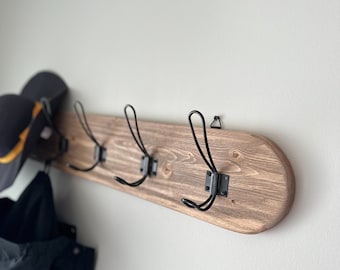 May include: A wooden coat rack with four black metal hooks. The rack is stained a dark brown color and has a slightly rounded shape. A black jacket and a black and yellow hat are hanging on the hooks.