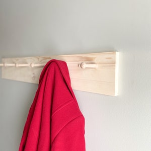 May include: A wooden coat rack with five hooks, painted white, mounted on a light gray wall. A red jacket is hanging on one of the hooks.