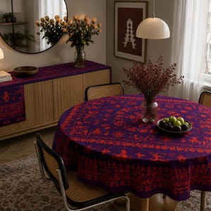May include: A round tablecloth featuring a deep purple base with a vibrant red intricate folk-style pattern. The design includes repeating motifs of stylized figures, animals, and floral elements arranged in concentric circular bands.
