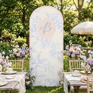 May include: A tall, arched event backdrop featuring a cream-colored surface with a light blue floral pattern and a central pink scalloped emblem displaying the letter B in a script font, positioned between two wooden tables with floral arrangements.