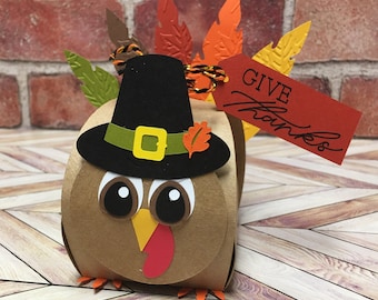 May include: A brown paper turkey-shaped box with a black pilgrim hat, googly eyes, and a red tag that says "GIVE Thanks". The turkey has colorful paper feathers and a red wattle. A festive Thanksgiving decoration.
