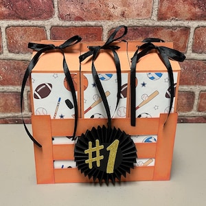 May include: Three orange boxes with black ribbon bows and sports themed designs. The boxes are arranged in a crate with a black and white ribbon with the number 