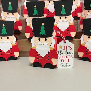 May include: A group of red and black nutcracker soldier papercraft ornaments with a white beard and gold accents. Each ornament has a green Christmas tree on the hat. A white tag with the text "It's Christmas so let's get crackin'" is attached to one of the ornaments.