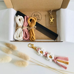 May include: A kit for making a macrame keychain with a daisy design. The kit includes three spools of cotton cord in white, pink, and yellow, a comb, a gold key ring, and a finished example of the keychain.