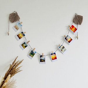 May include: A wall-mounted photo display with polaroid-style photos clipped to a string with wooden pegs. The photos show landscape scenes. The display is decorated with macrame and a vase of dried flowers. The wall is painted white.