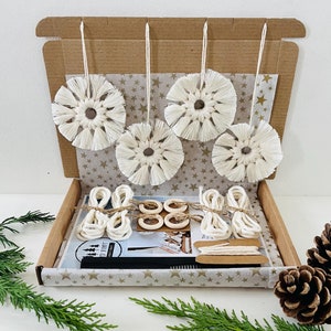 Op de afbeelding: Een doos gevuld met benodigdheden voor het maken van macramé sneeuwvlok ornamenten. De doos is open en toont vijf witte macramé sneeuwvlokken, houten ringen en wit touw. De doos is bekleed met gouden sterren op een zwarte achtergrond.