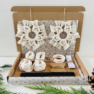 May include: A kit for making two macrame snowflake ornaments. The kit includes white cotton cord, wooden rings, a comb, a tape measure, and a small spool of twine. The kit is packaged in a brown cardboard box with a gold star pattern.