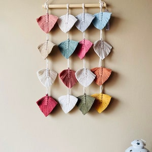 May include: A wall hanging made of macrame with a wooden dowel at the top. The macrame is in the shape of leaves and is in various colors, including pink, red, green, blue, yellow, and white.