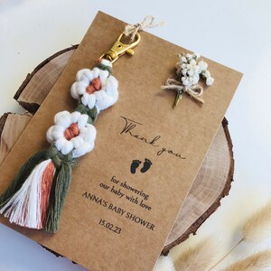 Peut inclure: Une carte de remerciement avec un petit bouquet de fleurs blanches et un porte-clés en macramé blanc et vert avec un anneau de clé doré. La carte dit "Merci d'avoir arrosé notre bébé d'amour, Baby Shower d'Anna, 15.02.23".