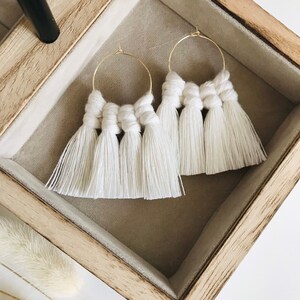 Puede incluir: Un par de pendientes de aro de macramé blanco con aros dorados. Los pendientes tienen un diseño de borla y se muestran en una caja de madera.