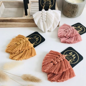 May include: Three pairs of handmade tassel earrings in white, mustard yellow, and terracotta. The earrings are made with cotton thread and have a layered, fan-shaped design. Each pair is displayed on a black card with the text "Wild Knit" and a small image of three trees.
