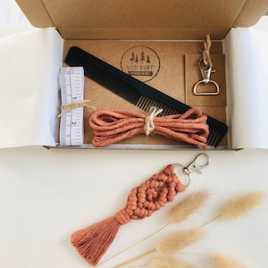 May include: A kit for making a macrame keychain. The kit includes a spool of brown cord, a black comb, a measuring tape, a silver key ring, and a tag with the text "WILD KNOT" in a circle.