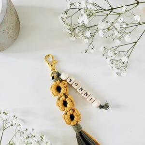 May include: A macrame keyring featuring three mustard yellow flower-shaped knots, a gold-toned metal lobster clasp, and a multi-coloured tassel in olive green and yellow. A strand of light wooden beads spells out the name SONNI in black letters.