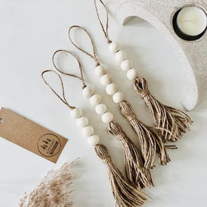 May include: Four white wooden bead garlands with jute tassels. Each garland has a loop at the top for hanging. The garlands are on a white surface with a brown plant in the background.