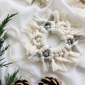 Peut inclure: Une d&eacute;coration en macram&eacute; blanche avec des accents argent&eacute;s, pr&eacute;sentant un motif circulaire avec des d&eacute;tails en frange. L'ornement est pr&eacute;sent&eacute; sur un fond en tissu blanc.