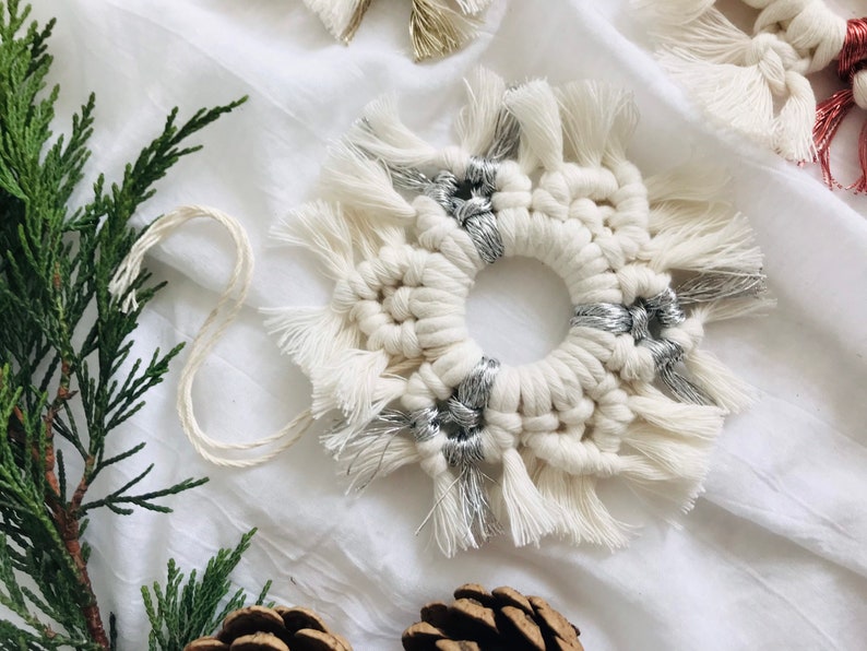 Peut inclure: Une d&eacute;coration en macram&eacute; blanche avec des accents argent&eacute;s, pr&eacute;sentant un motif circulaire avec des d&eacute;tails en frange. L'ornement est pr&eacute;sent&eacute; sur un fond en tissu blanc.
