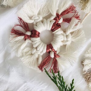 Peut inclure: Une d&eacute;coration de flocon de neige en macram&eacute; blanc et cuivre avec un anneau au centre. La d&eacute;coration est sur un fond de tissu blanc avec une brindille verte.