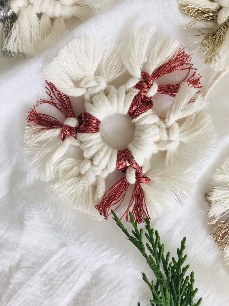Peut inclure: Une d&eacute;coration de flocon de neige en macram&eacute; blanc et cuivre avec un anneau au centre. La d&eacute;coration est sur un fond de tissu blanc avec une brindille verte.