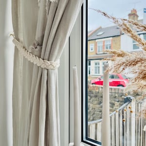 May include: Beige linen curtains with a braided rope tieback. Two white taper candles sit in decorative holders on a windowsill. The window overlooks a residential street with a red car.