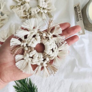 Peut inclure: Une d&eacute;coration de flocon de neige en macram&eacute; blanc et or avec un centre circulaire. L'ornement est tenu dans une main sur un fond blanc.