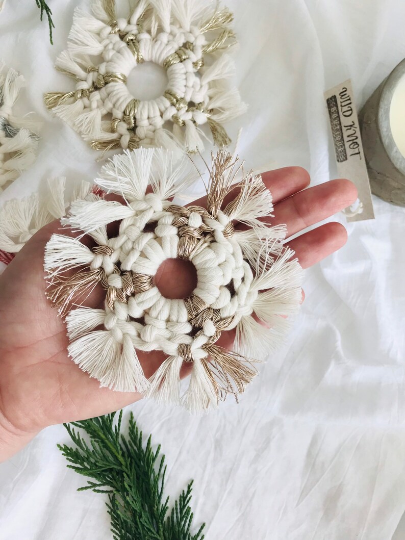 Peut inclure: Une d&eacute;coration de flocon de neige en macram&eacute; blanc et or avec un centre circulaire. L'ornement est tenu dans une main sur un fond blanc.