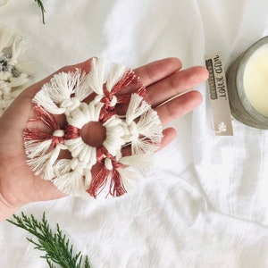 Peut inclure: Une d&eacute;coration de No&euml;l en macram&eacute; en forme de flocon de neige, de couleur blanche et cuivre, avec des franges. L'ornement est tenu dans une main avec une &eacute;tiquette qui indique "Wild Knot" en lettres noires.