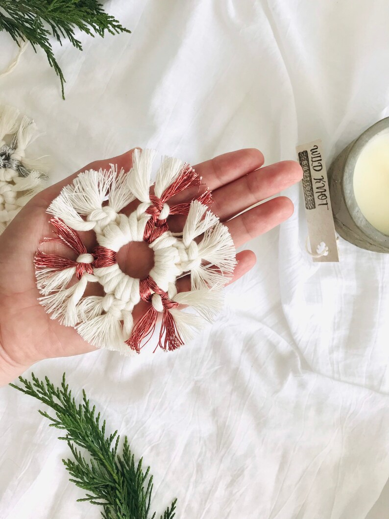 Peut inclure: Une d&eacute;coration de No&euml;l en macram&eacute; en forme de flocon de neige, de couleur blanche et cuivre, avec des franges. L'ornement est tenu dans une main avec une &eacute;tiquette qui indique "Wild Knot" en lettres noires.