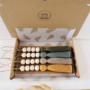 May include: Set of four wooden bead and tassel ornaments in a cardboard box. The tassels are in shades of green, blue, beige, and yellow. The box is lined with white paper with a gold fern pattern. The box has a logo that says "Wild & Free".