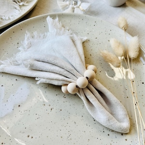 Op de afbeelding: Een beige linnen servet met een gerafelde rand, vastgehouden door een houten kralen servetring, rust op een gespikkelde keramische plaat. De setting omvat een tweede bord, een wijnglas en decoratieve droogbloemen.