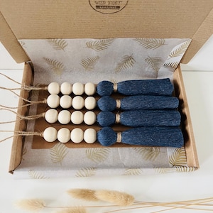 Può includere: Set di sei appendini da parete con perline di legno e nappe blu in una scatola di cartone con carta velina a motivi di foglie dorate. La scatola ha un logo che dice "Wild Nest" con un design ad albero.