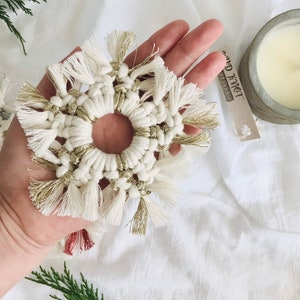 Peut inclure: Une d&eacute;coration de No&euml;l en macram&eacute; en forme de flocon de neige blanc et or avec un trou au centre. L'ornement est tenu dans une main sur un fond blanc.