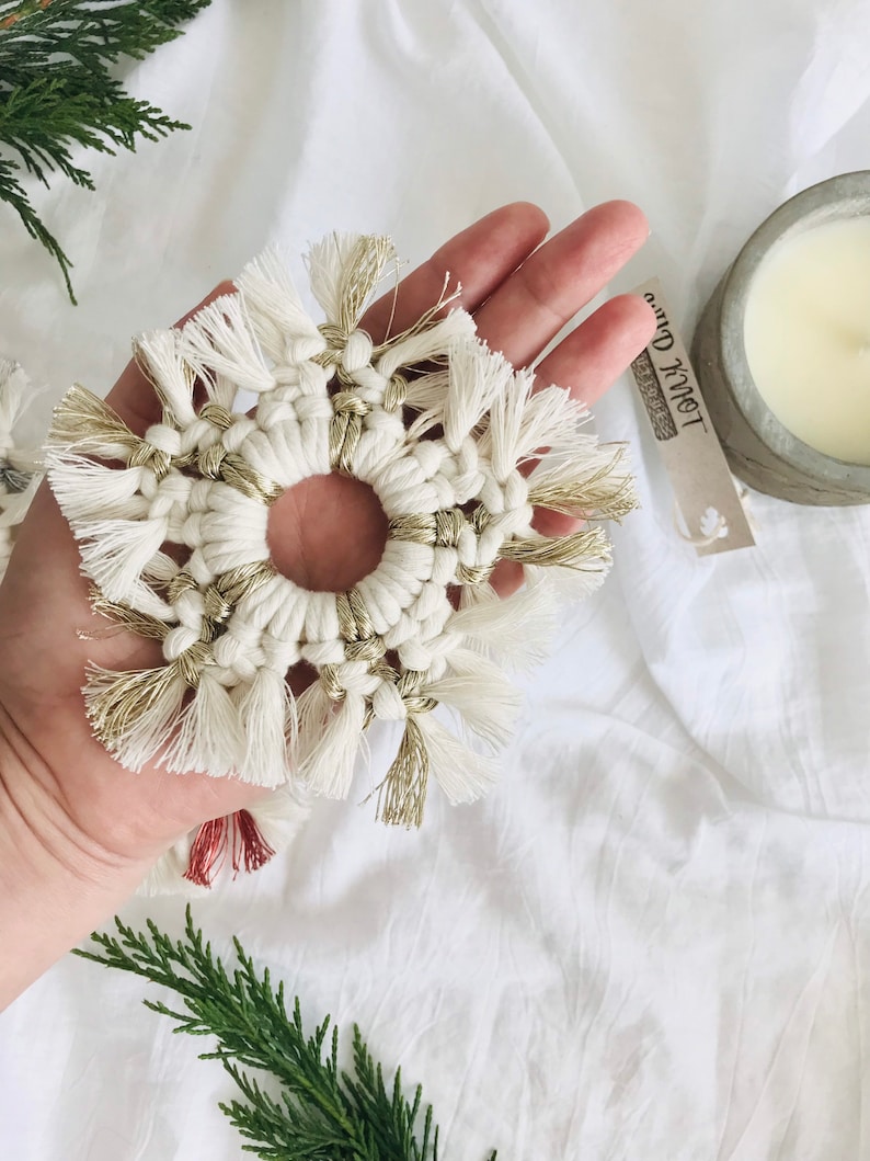 Peut inclure: Une d&eacute;coration de No&euml;l en macram&eacute; en forme de flocon de neige blanc et or avec un trou au centre. L'ornement est tenu dans une main sur un fond blanc.