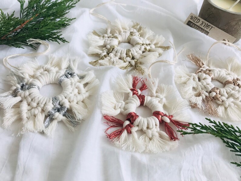 Peut inclure: Quatre ornements en macram&eacute; faits &agrave; la main en forme de flocons de neige avec des accents de diff&eacute;rentes couleurs. Les ornements sont blancs avec des accents argent&eacute;s, dor&eacute;s et rouges. Les ornements sont suspendus &agrave; un fond blanc.