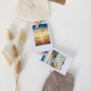 May include: A white and beige macrame tassel garland with a wooden ring at the top. The garland has a series of tassels and photo clips, with two instant photos of a beach scene attached.