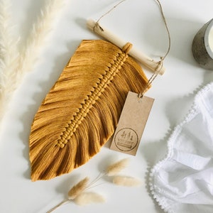 May include: A mustard yellow macrame feather wall hanging with a wooden dowel and a brown paper tag with the text "Wild Knot" and a logo of three trees.