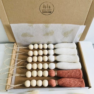 Peut inclure: Un ensemble de guirlandes de perles en bois avec des pompons dans une boîte en carton. Les guirlandes sont faites de perles en bois naturel et ont des pompons blancs et terracotta. La boîte est décorée d'un motif de feuilles et les mots "Wild Knot" sont imprimés sur le dessus.