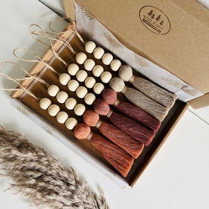 Peut inclure: Un ensemble de six guirlandes de perles en bois avec des pompons dans des tons de brun, beige et crème. Les guirlandes sont emballées dans une boîte brune avec un papier de soie blanc et un logo qui dit "wild & free".