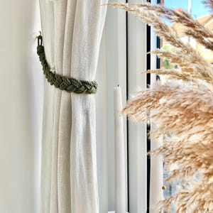Peut inclure: Un embrasse-rideau macramé vert olive tressé maintient un rideau crème. Le macramé est fait de corde épaisse. Une bougie blanche est dans un support blanc. De l'herbe de pampa séchée est en arrière-plan.