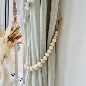 May include: A set of wooden beads strung together to create a curtain tieback. The beads are a light natural wood colour and are strung on a natural twine cord. The tieback is hanging on a light grey linen curtain.