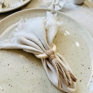 May include: A cream linen napkin with frayed edges, secured by a wooden ring and tied with twine, sits on a speckled ceramic plate. The scene suggests a rustic, elegant dining theme. Another plate and a wine glass are also visible.