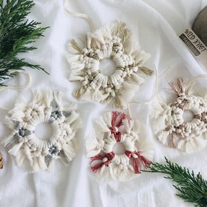 Peut inclure: Quatre ornements en macram&eacute; faits &agrave; la main en forme de flocons de neige avec un anneau au centre. Les ornements sont blancs avec des accents argent&eacute;s, dor&eacute;s et rouges. Les ornements sont suspendus &agrave; un fond en tissu blanc.
