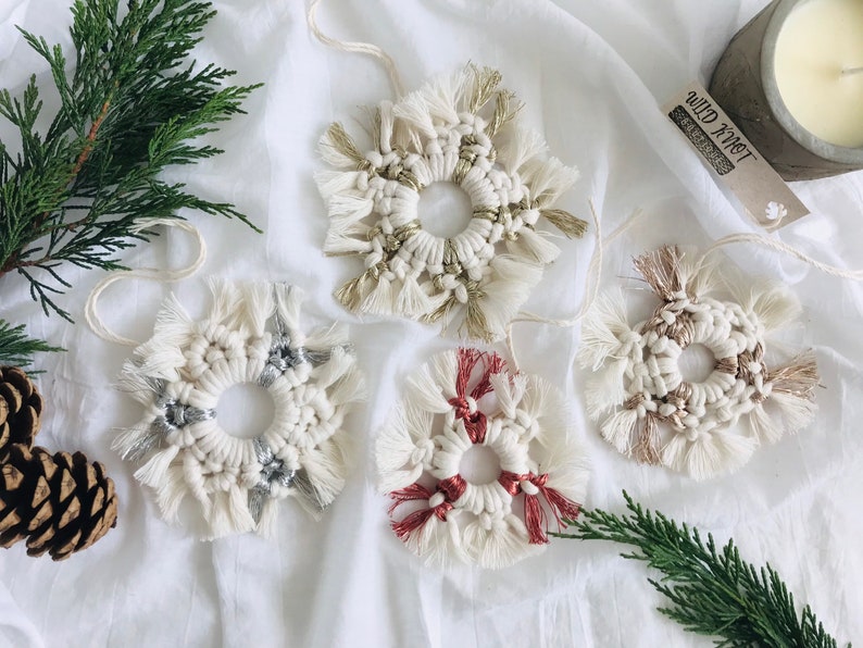 Peut inclure: Quatre ornements en macram&eacute; faits &agrave; la main en forme de flocons de neige avec un anneau au centre. Les ornements sont blancs avec des accents argent&eacute;s, dor&eacute;s et rouges. Les ornements sont suspendus &agrave; un fond en tissu blanc.