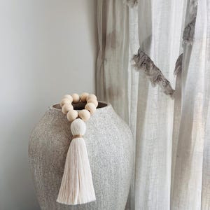 May include: A decorative loop of smooth, light-coloured wooden beads featuring a long, off-white fibre tassel tied with a thin cord. The beaded accessory is draped over the rim of a large, textured, light grey ceramic vase with a tapered shape.