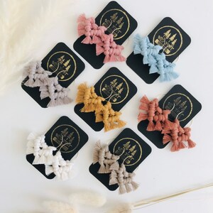 May include: A set of 10 pairs of handmade macrame earrings in various colours, including beige, white, light blue, light pink, and mustard yellow. Each pair features a geometric design with a gold hoop and a tassel fringe. The earrings are displayed on black cards with the brand name "Wild West" printed on them.